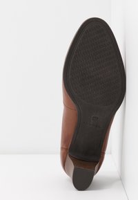 Chaussure en cuir marron au design plat, avec une tige en matériau lisse et une semelle extérieure en caoutchouc noir texturée. Elle présente des détails de coutures discrètes.