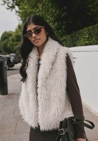 Kunstpelzweste in Elfenbein mit einer zotteligen Textur, ausgestattet mit einem breiten Kragen. Trägt sie über einem dunklen Kleid, kombiniert mit rechteckigen Sonnenbrillen und einer schwarzen Tasche.