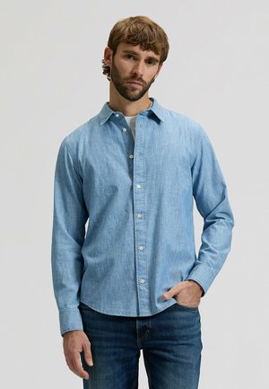 Mężczyzna o brązowych włosach i brodzie, ubrany w jasnoniebieską koszulę zapinaną na guziki na białą podkoszulkę oraz ciemnoniebieskie dżinsy, stojący na jednolitym tle.