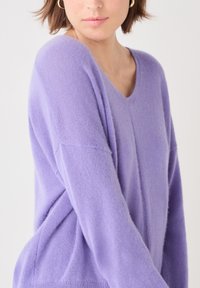 Lila v-ringad tröja gjord av mjukt, fuzzy tyg med sänkt axel och avslappnad passform. Har subtila ribbade detaljer vid nederkanten.