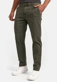 Pantalones de algodón verde oliva con corte recto, cinco bolsillos y detalle de logo bordado. Combinados con zapatillas blancas.