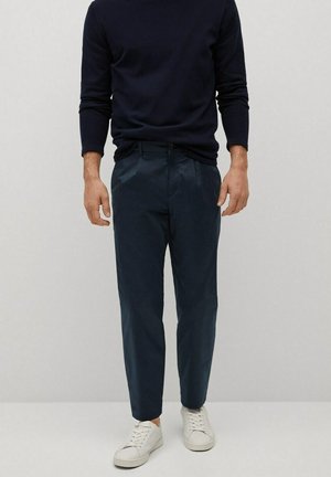 Homme portant un pull marine foncé, un pantalon bleu foncé et des baskets blanches, debout devant un fond clair uni.