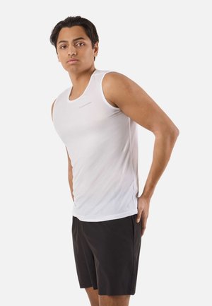 Giovane uomo che indossa una canotta bianca senza maniche e pantaloncini neri, in piedi con una mano posata sui fianchi.