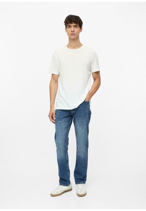 Jonge man staat met één hand in zijn zak en draagt een effen wit t-shirt, blauwe spijkerbroek en witte sneakers tegen een witte achtergrond.