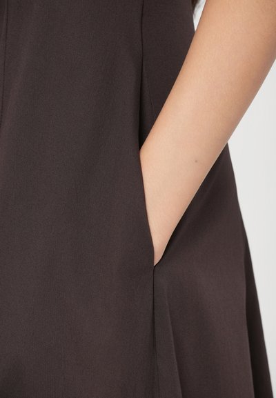 Tmavohnedá látka na šaty s matnou textúrou, s bočnou kapsou a elegantným strihom. Detailný záber ruky odpočívajúcej blízko kapsy.