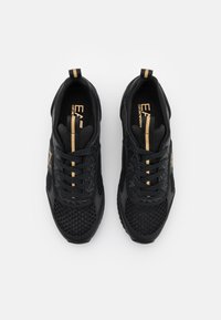 Zapatillas deportivas negras con parte superior de malla, suela de goma, cordones negros y detalles en dorado. Presentan una pestaña para tirar y un diseño estampado en el lateral.