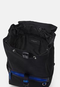 Sac à dos en nylon noir avec fermeture à cordon, intérieur doublé avec une étiquette logo, et une poche zippée bleue sur le devant.