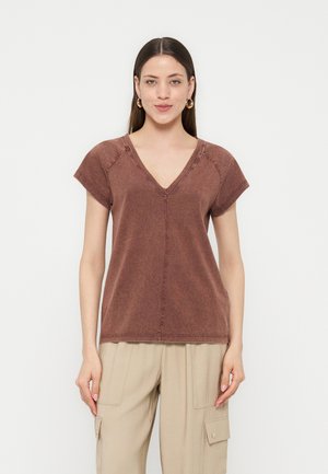 Γυναίκα που φοράει καφέ μπλουζάκι με V λαιμόκοψη και κοντά μανίκια και μπεζ παντελόνι cargo, στέκεται μπροστά σε απλό λευκό φόντο.