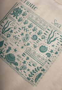 Tejido de color beige claro con un diseño étnico en tonos azul verdoso que presenta conchas marinas, peces, algas y una tortuga, enmarcado por bordes decorativos.