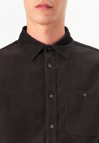 Camisa de pana marrón con cuello abotonado, bolsillo frontal y seis botones, que presenta un patrón de tejido texturizado y un brillo sutil.