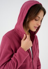 Felpa con cappuccio in micropile di color rosa scuro, dotata di chiusura a zip e un cappuccio ampio. Caratterizzata da un tessuto testurizzato e polsini aderenti.