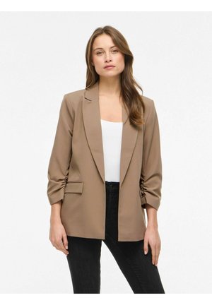 Femme aux longs cheveux bruns portant un blazer beige à manches froncées sur un haut blanc et un jean noir, debout devant un fond blanc.