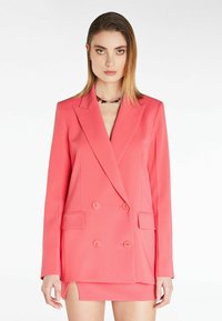 Blazer doppiopetto corallo con rever a punta, tasche frontali e bottoni decorativi. Tessuto liscia, design strutturato, abbinato a una mini gonna.