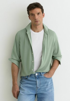 Mand iført en lysegrøn skjorte med knapper over en hvid t-shirt og lyseblå jeans, stående mod en hvid væg med den ene hånd i lommen.