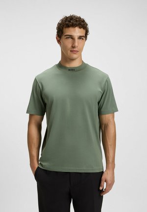 Uomo con capelli ricci che indossa una tshirt a maniche corte verde chiaro con "BOSS" sul colletto e pantaloni neri su uno sfondo semplice.