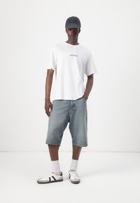 T-shirt bianco a maniche corte con testo nero, pantaloni corti di denim blu e sneakers bianche con strisce nere. Un cappellino grigio completa l'outfit.