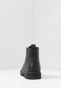 Botín de cuero negro hasta el tobillo con un acabado texturizado, punta redonda, tacón bajo y etiqueta de logo roja en el lateral. Diseño minimalista con líneas limpias.