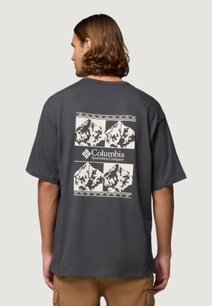 Persona con capelli di lunghezza media vista di spalle, indossa una maglietta grigio scuro con grafiche di montagne bianche e la scritta "Columbia Sportswear Company".