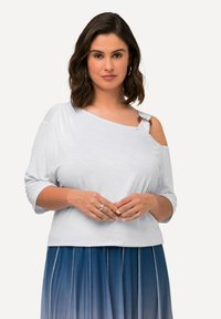 Top azul claro, con los hombros al descubierto y un acento de botón en un lado, con mangas cortas y un ajuste relajado. Textura suave.