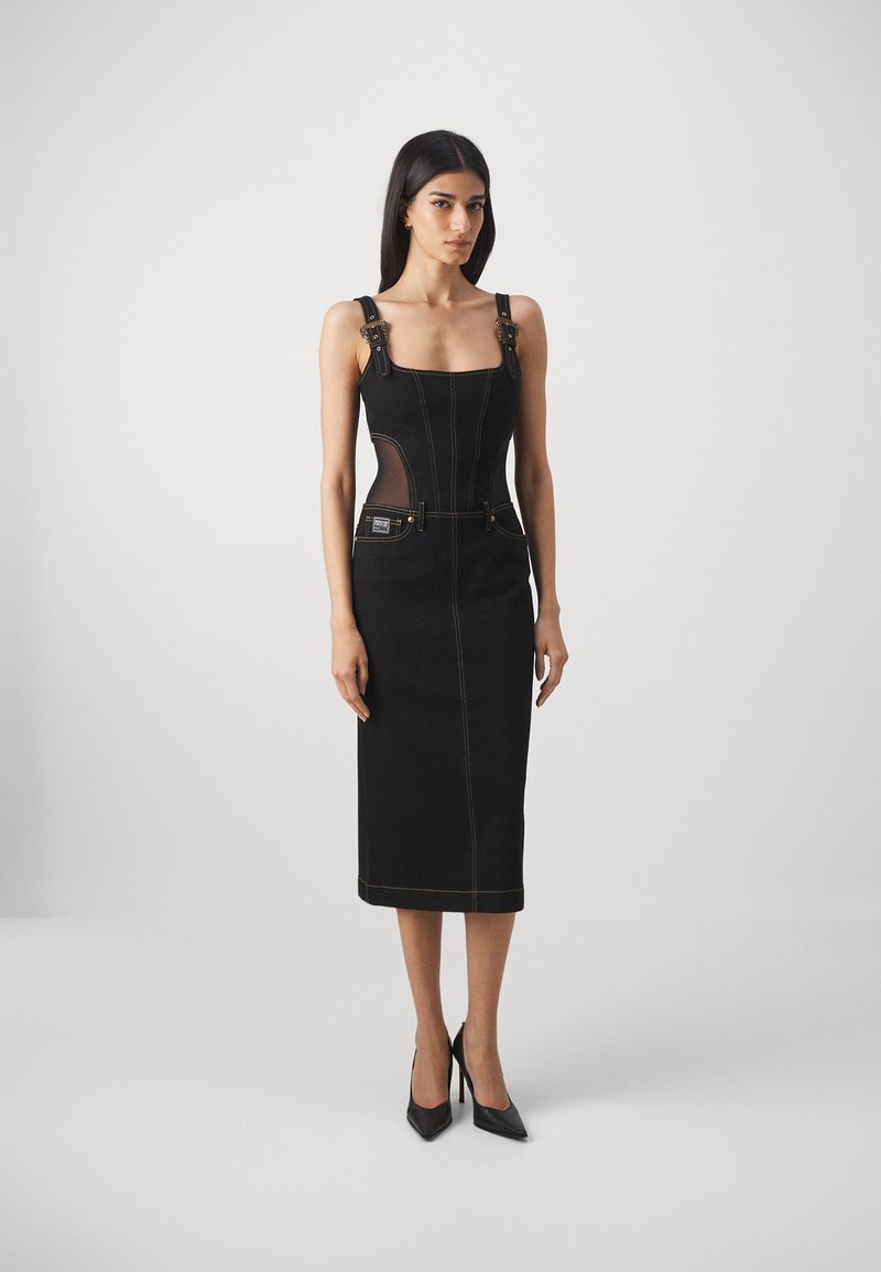 Femme portant une robe midi noire ajustée avec des bretelles à boucle, des panneaux latéraux en maille, des coutures apparentes, des poches avant et des talons hauts noirs à pointes.