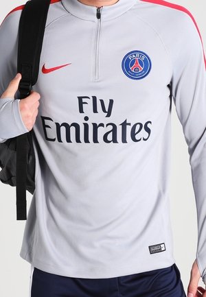 Person trägt ein graues Paris Saint-Germain Trainingstrikot mit Fly Emirates Logo und trägt einen schwarzen Rucksack auf einer Schulter.