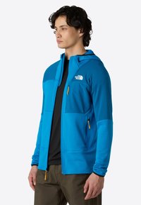 Giacca blu con zip e cappuccio, realizzata in materiale testurizzato, con una tasca sul petto e dettagli gialli sulla zip. Indossata sopra una maglietta nera.