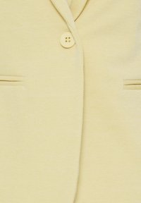 Gros plan sur le devant d'un blazer jaune pâle avec un bouton assorti unique et deux poches horizontales fendues.