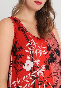Rotes ärmelloses Oberteil mit floralen und botanischen Mustern in Schwarz und Weiß, ergänzt durch ein subtil gestreiftes Design. Hergestellt aus leichtem Stoff.