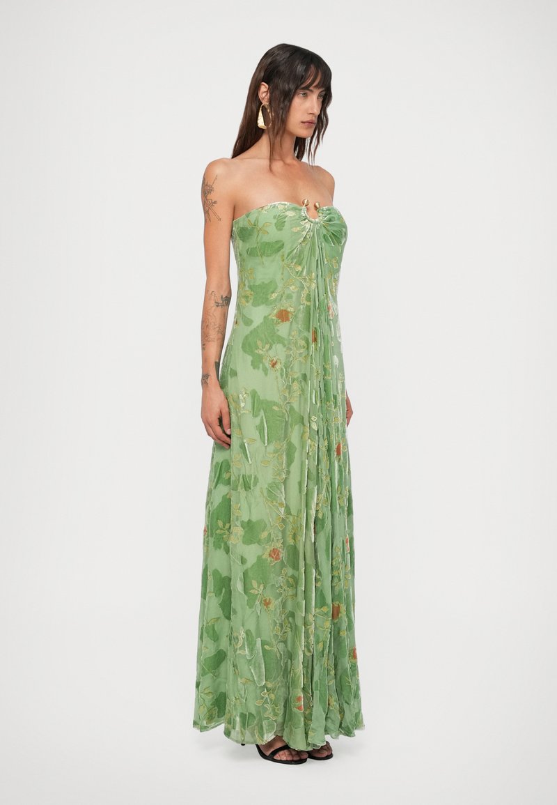 Woman with long dark hair and tattoos wearing strapless green floral maxi dress and black sandals, standing against white background.