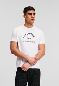 Balta trumpomis rankovėmis marškinėliai, pagaminti iš medvilnės, su juodu grafiniu spaudiniu, ant kurio užrašyta "KARL LAGERFELD PARIS", ir apvaliu iškirpimu.