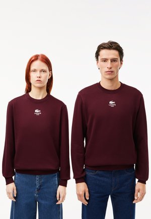 Sudaderas de color burdeos en tejido suave, con cuello redondo y puños acanalados, que lucen un logo blanco de Lacoste en el pecho. Se llevan con jeans azules.