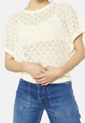 Kvinde iført en cremefarvet, kortærmet, strikket top med en bølget kant og blå denimjeans, hænderne samlet ved taljen.