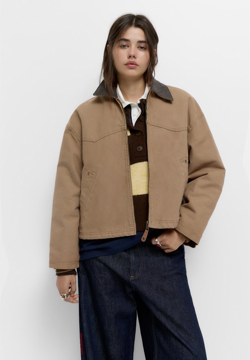 Brun canvasjakke med en mørkere læderkrave, frontlynlåslukning, to lommer, båret over en brun og stribet sweater. Mørke jeans synlige.