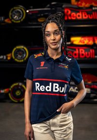 Ciemnoniebieska koszulka polo z czerwonymi akcentami i brandingiem 'Red Bull', z logo oraz kołnierzykiem. W tle znajdują się samochody wyścigowe.