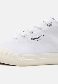 Valkoinen matala kangaskenkä valkoisilla nauhoilla ja kumipohjalla, jossa on "Pepe Jeans London" -logo sivulla.