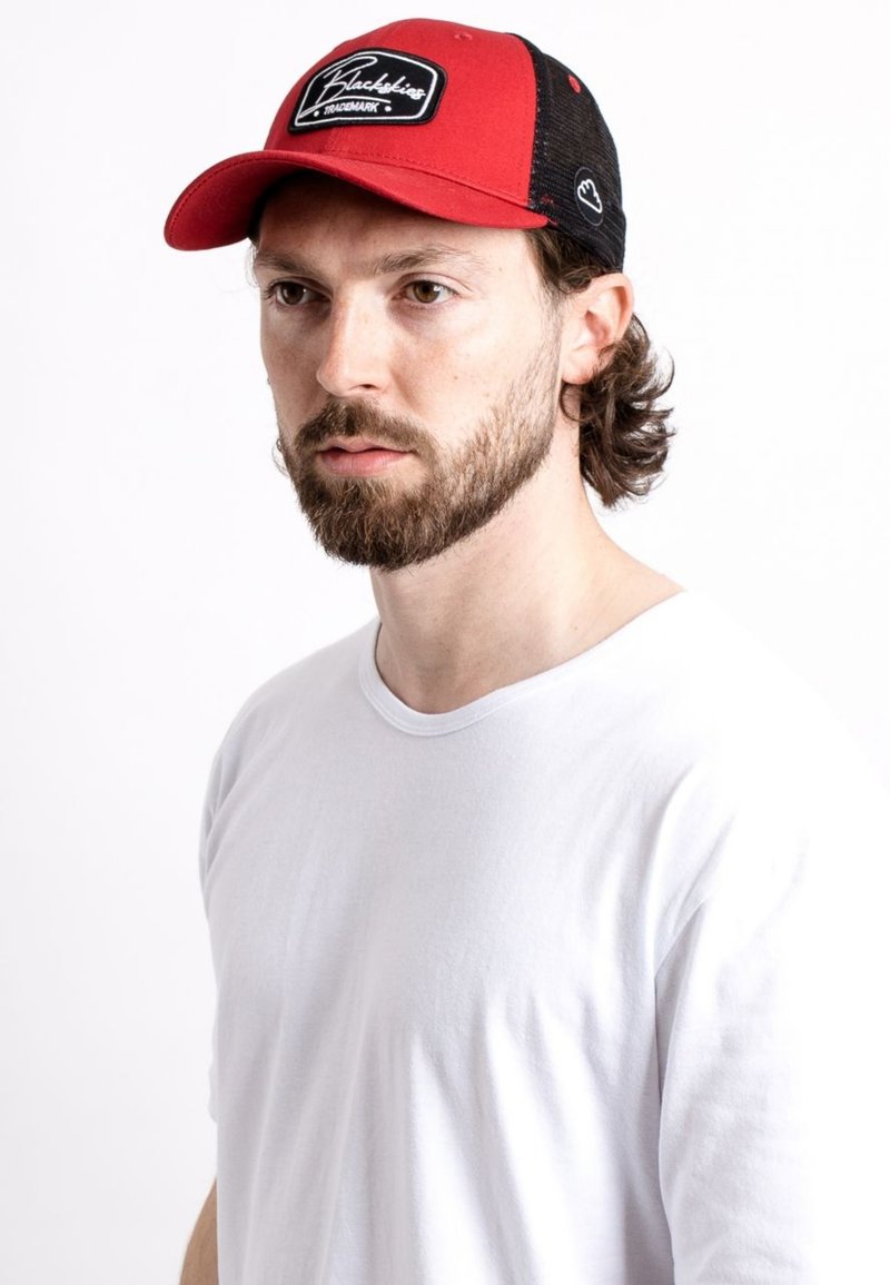 Cappellino rosso e nero con parte posteriore in rete, dotato di un patch rettangolare con logo. Chi lo indossa ha una maglietta bianca semplice e una leggera barba.
