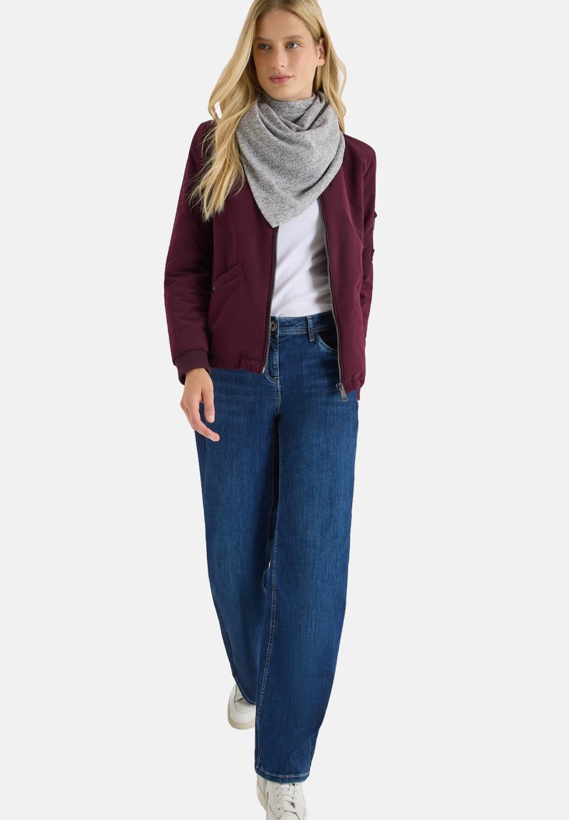 Kvinde går iført hvide sneakers, brede blå jeans, hvid top, maroon jakke og grå tørklæde om halsen på hvid baggrund.