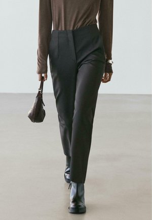 Pantaloni neri su misura con vita strutturata, abbinati a un top marrone a maniche lunghe e stivaletti neri, con una piccola borsa in mano.