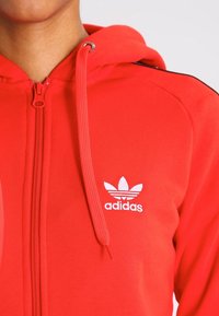 Rode zip-up hoodie van zacht materiaal, met een wit Adidas-logo op de linker borst en een koord met metalen punten.