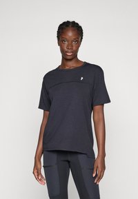 Kvinde med fletning iført en løs mørkegrå t-shirt og mørke leggings med lommer, stående mod en ensfarvet lys baggrund.