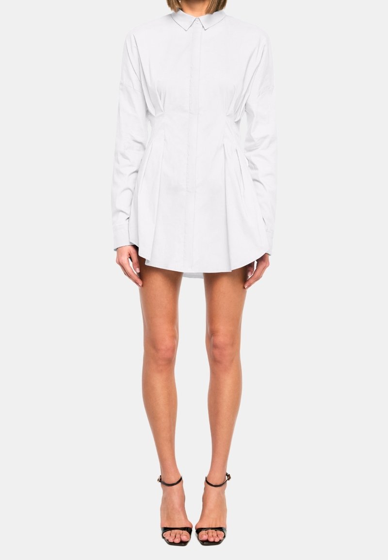 Femme portant une robe-chemise blanche à manches longues avec des plis, associée à des sandales noires à talons hauts et bride à la cheville, debout sur fond blanc.