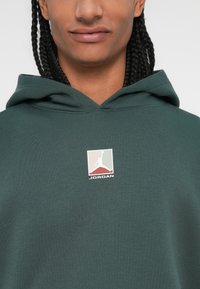 Tmavo zelená mikina s klokankou. Na hrudi sa nachádza malé biele a farebné logo Jordan. Tkanina vyzerá na dotyk mäkko a textúrovo.