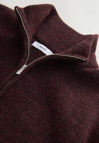 Gros plan d'un pull en maille duveteuse bordeaux avec une demi-fermeture éclair argentée et une étiquette en tissu blanc portant la mention "NEXT" à l'intérieur du col.