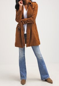 Brązowy zamszowy płaszcz trench z zapięciem w talii, skośnymi kieszeniami i detalami guzików, w połączeniu z dzwonami w jasnoniebieskim kolorze oraz butami w panterkę.