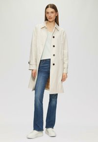Beige-Trenchcoat mit großen schwarzen Knöpfen, getragen über einem hellblauen Oberteil. Kombiniert mit blauen Jeans und weißen Sneakers. Glatte Textur, leicht lockere Passform.