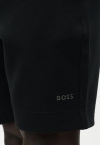 Κοντινό πλάνο μαύρων σορτς με κεντημένο λογότυπο "BOSS" κοντά στο τελείωμα στο αριστερό πόδι, φορεμένα από άτομο με σκουρόχρωμο δέρμα.