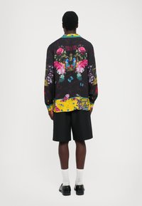 Bărbat îmbrăcat cu pantaloni scurți negri și o cămașă cu mâneci lungi, împodobită cu desene colorate de flori, fluturi și coroane, stând cu spatele împotriva unui fundal simplu.
