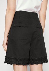Schwarze Shorts mit hoher Taille, versehen mit einer Spitzenborte am Saum und zwei aufgesetzten Taschen auf der Rückseite. Hergestellt aus einem glatten Stoff.