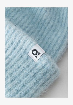 Helle blaue Strickmütze mit geripptem Muster. Verfügt über ein weißes Etikett mit einem schwarzen Logo, das die Designdetails akzentuiert.