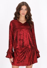 Rød, skinnende, fløjlsagtig kjole med lange ærmer. V-halsudskæring og flæsekantede manchetter. Draperet stil med løs pasform og tekstureret stof.